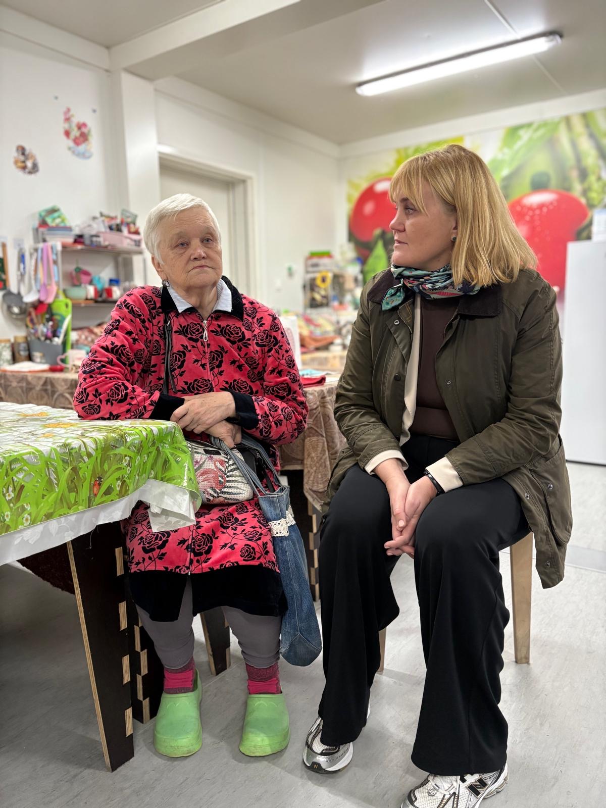 En eldre kvinne og en yngre kvinne sitter ved et bord i et rom med kjøkkenutstyr og matvarer i bakgrunnen.