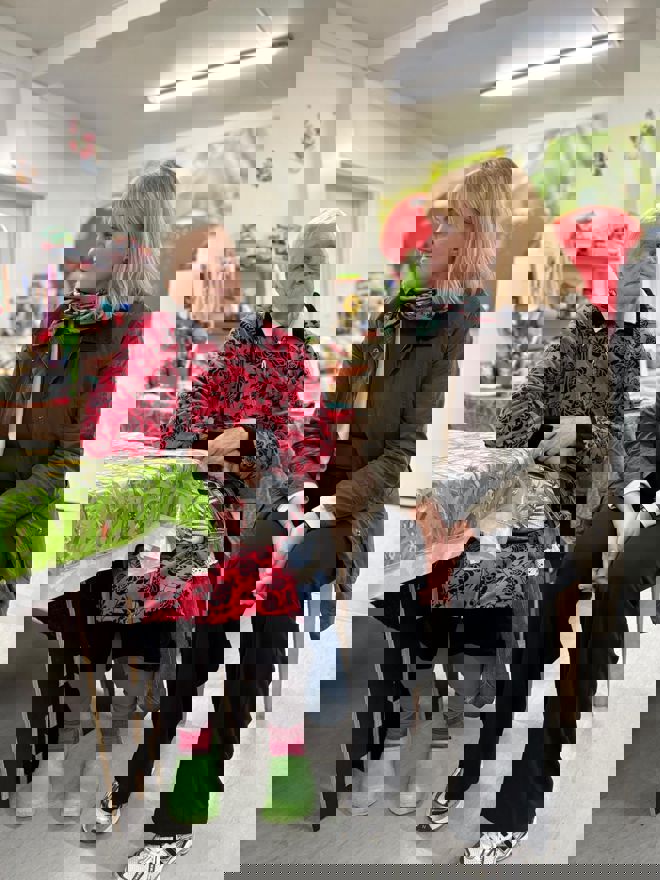 En eldre kvinne og en yngre kvinne sitter ved et bord i et rom med kjøkkenutstyr og matvarer i bakgrunnen.