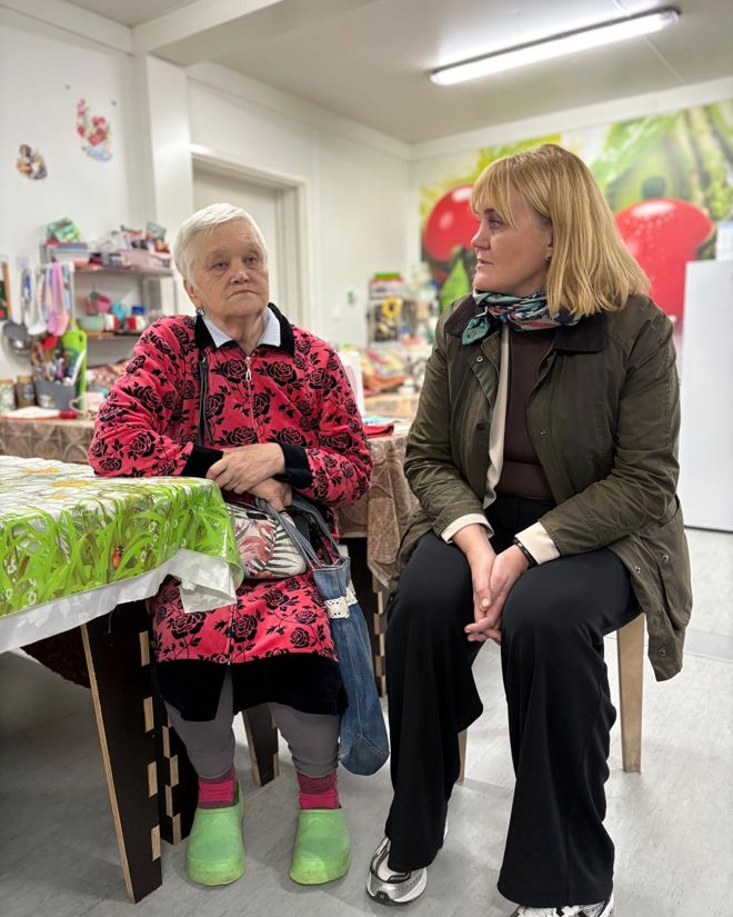 En eldre kvinne og en yngre kvinne sitter ved et bord i et rom med kjøkkenutstyr og matvarer i bakgrunnen.