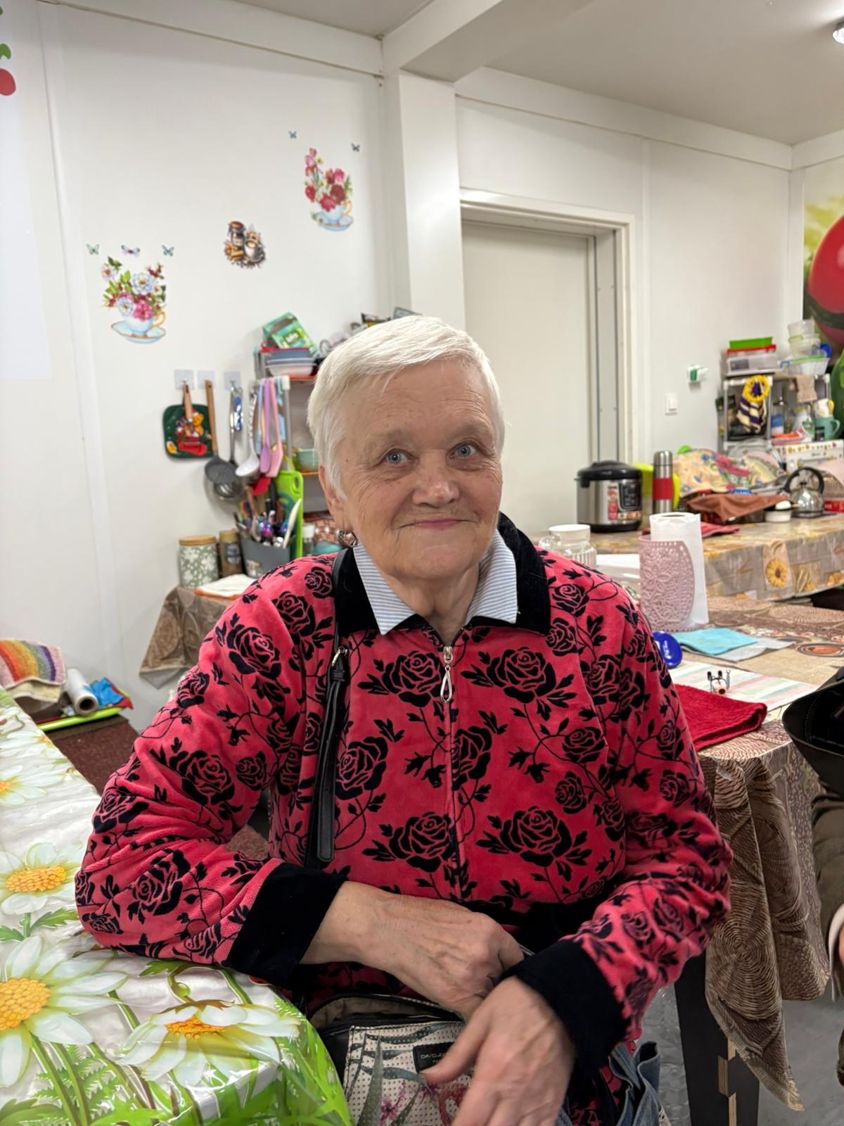 En eldre kvinne sitter ved et bord i et kjøkken eller oppholdsrom med kjøkkenutstyr og dekorasjoner i bakgrunnen.