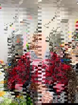 En eldre kvinne sitter ved et bord i et kjøkken eller oppholdsrom med kjøkkenutstyr og dekorasjoner i bakgrunnen.