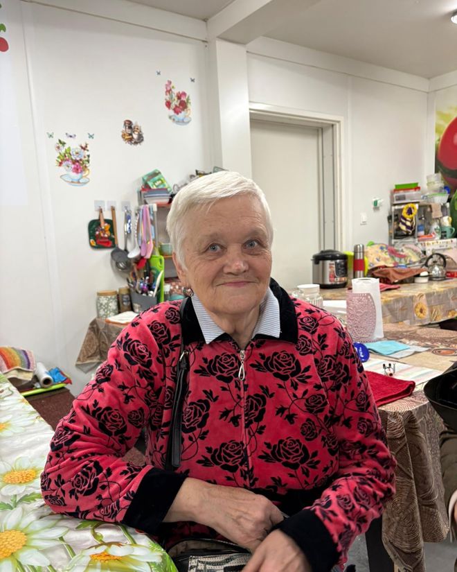 En eldre kvinne sitter ved et bord i et kjøkken eller oppholdsrom med kjøkkenutstyr og dekorasjoner i bakgrunnen.