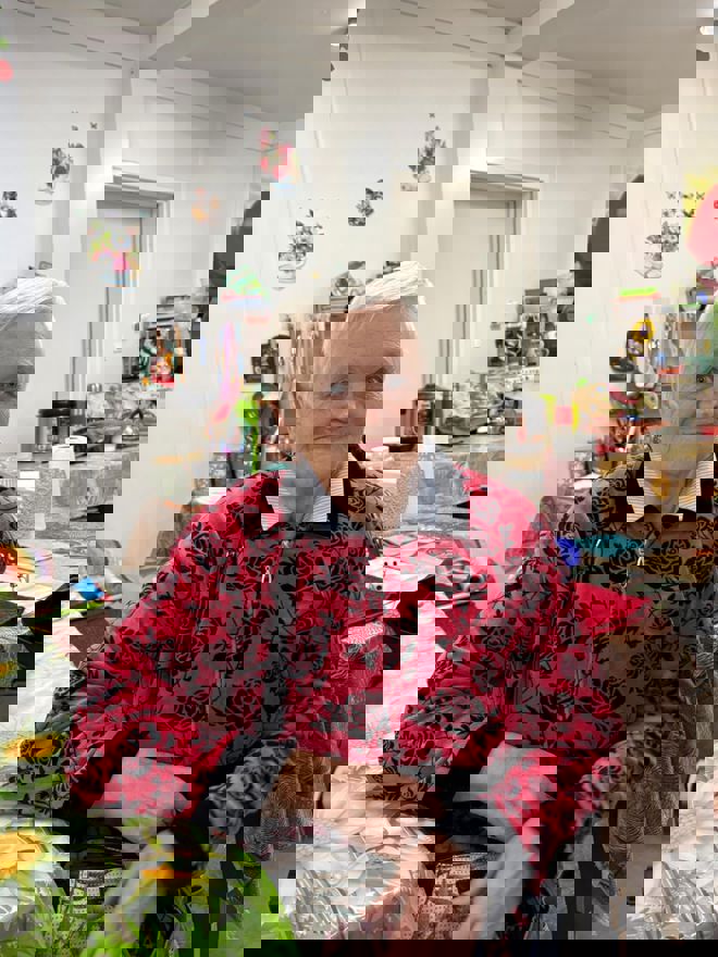 En eldre kvinne sitter ved et bord i et kjøkken eller oppholdsrom med kjøkkenutstyr og dekorasjoner i bakgrunnen.