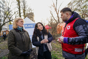 Three adults, two women and one man, are standing and talking outdoors in front of a tent with informational banners.