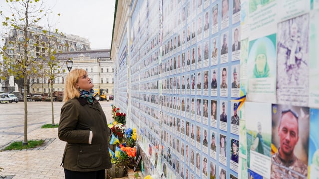 En kvinne står foran en minnevegg med bilder og navn, med blomster og ukrainske flagg plassert ved veggen.