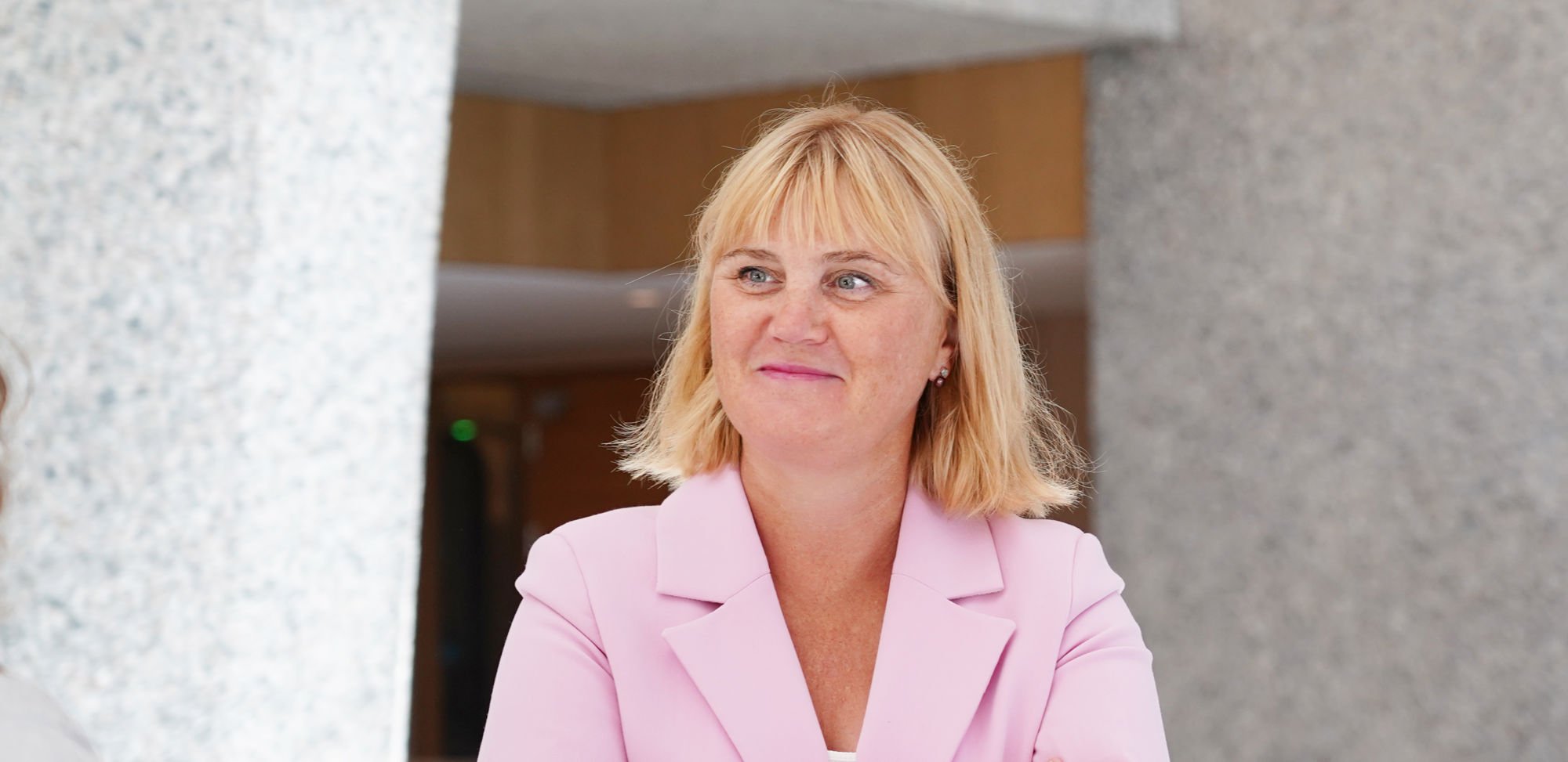 Bildet viser en kvinne med skulderlangt blond hår som står med armene i kors. Vedkommende har på seg en lyserosa dressjakke over en lys topp.