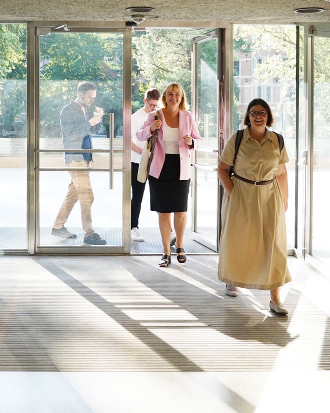 Bildet viser fire personer som er i ferd med å gå inn gjennom store glassdører til en lys og moderne bygning. Den nærmeste personen til høyre har på seg en lys, lang kjole og bærer en svart veske over skulderen. Litt lenger bak i midten ser man en person med blondt, skulderlangt hår, som har på seg en rosa blazer, hvit topp og svart skjørt