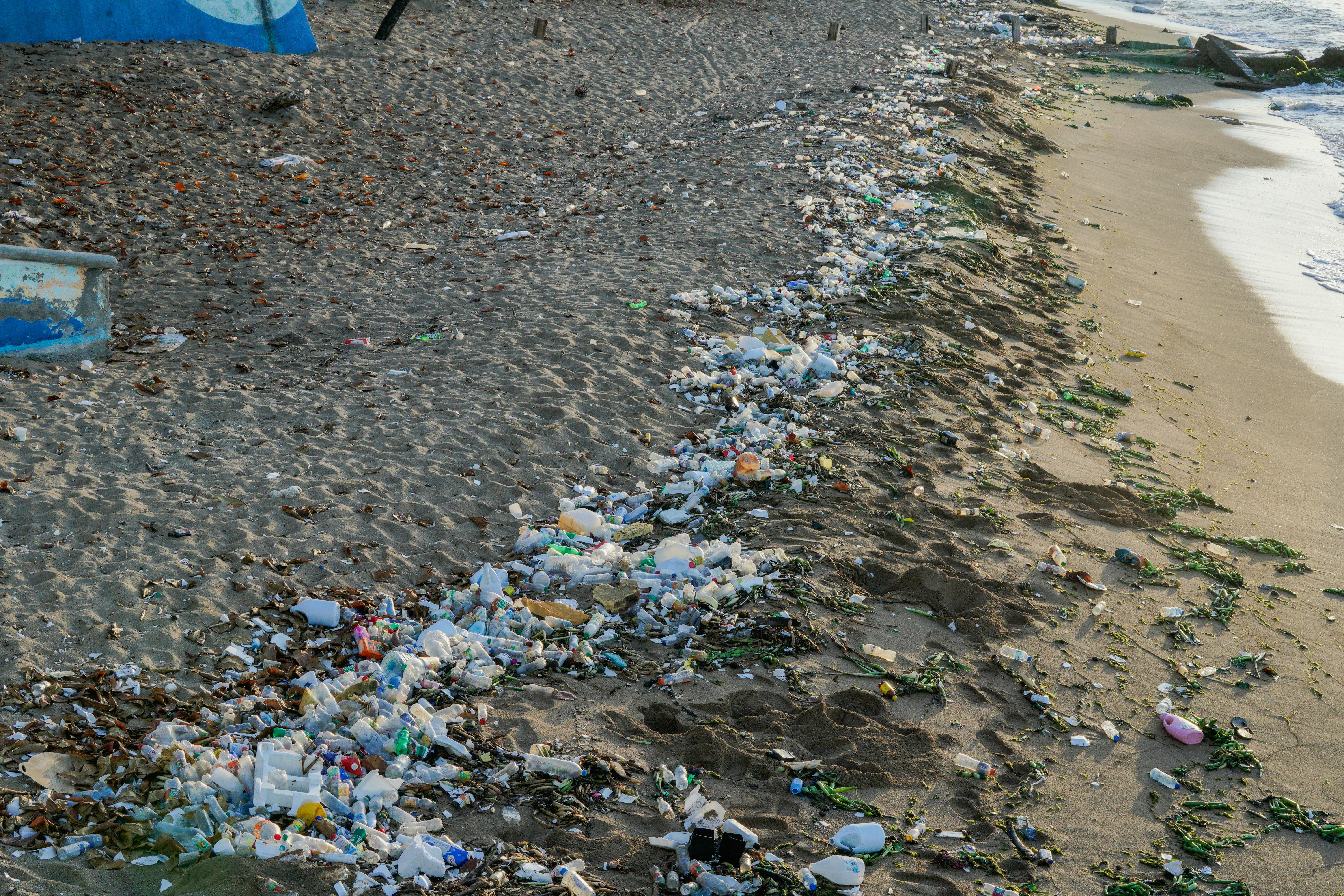 The image depicts a sandy beach heavily littered with trash and debris. A long line of garbage, including plastic bottles, food wrappers, and other waste, stretches along the shoreline, mixed with seaweed and natural materials. 
