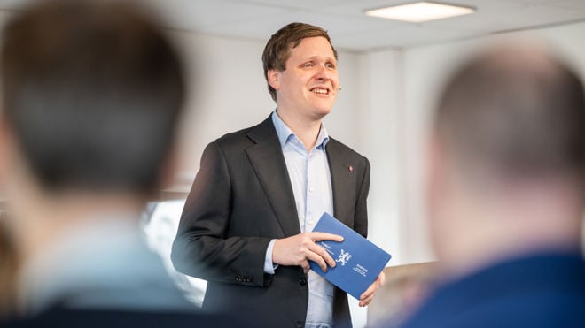 Bildet viser utviklingsminister Åsmund Grøver Aukrust som står i et rom med hvite vegger og tak, sannsynligvis i en formell eller profesjonell setting. Personen er kledd i en mørk dressjakke med en lys skjorte under. I den ene hånden holder personen en blå mappe med det norske riksvåpenet trykt på forsiden