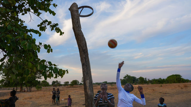 This image captures a lively outdoor scene where children are playing basketball in a rural, open field. The basketball hoop is quite simple—it’s made from a rough wooden post with a metal ring attached at the top, instead of a traditional backboard. Two children are reaching up enthusiastically, either trying to catch or shoot a basketball that is in mid-air above them. The ground is dry and dusty, with a few scattered trees providing greenery. In the background, more children can be seen, some standing and one with a bicycle, watching or waiting their turn. The sky is wide and open, with soft clouds and the warm light of either early morning or late afternoon.