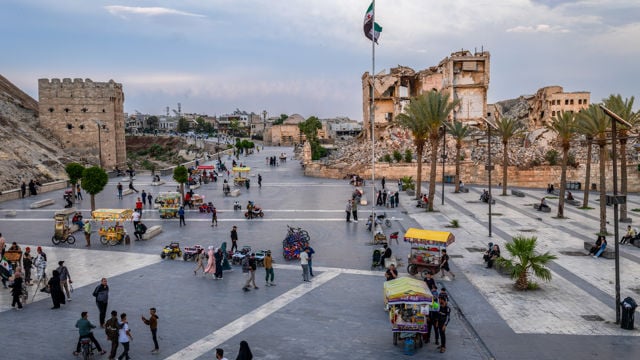 Et stort åpent torg med mange mennesker, flere boder som selger varer, palmetrær og en stor bygning med skader i bakgrunnen. Et stort nasjonalflagg vaier på en høy flaggstang.