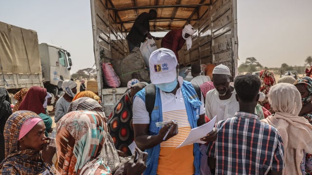 Bildet viser en humanitær hjelpearbeider fra UNHCR som registrerer informasjon fra en gruppe mennesker samlet rundt en lastebil. Lastebilen er lastet med forsyninger, og folkene er kledd i fargerike klær og hodeplagg. Scenen skjer utendørs i et tørt område.