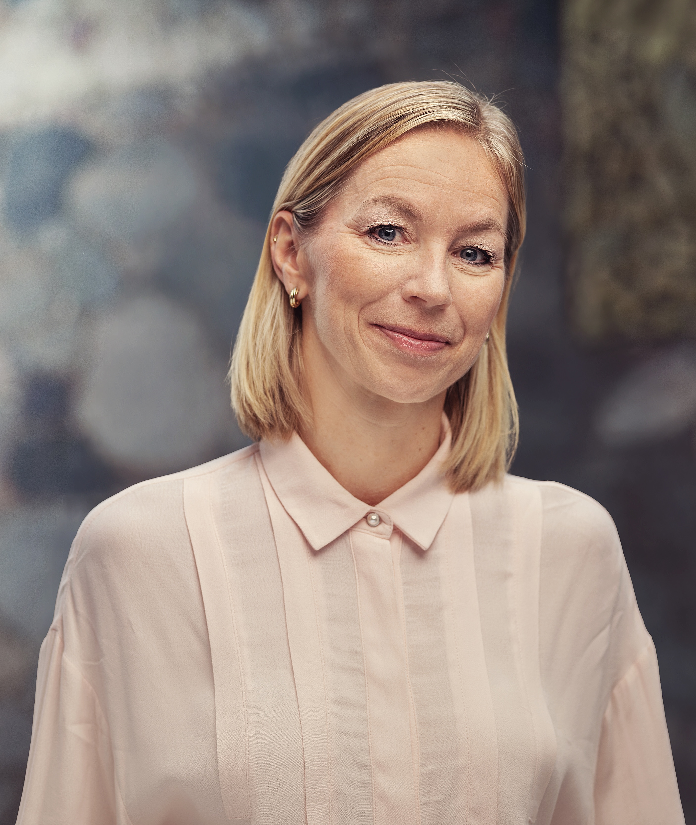 Et portrett av Monica Svenskerud. Hun står med skulderlangt blondt hår og har på seg en lys rosa skjorte med krage. Bakgrunnen er mykt uskarp med duse blå og grå toner, og gir bildet en rolig og nøytral stemning.