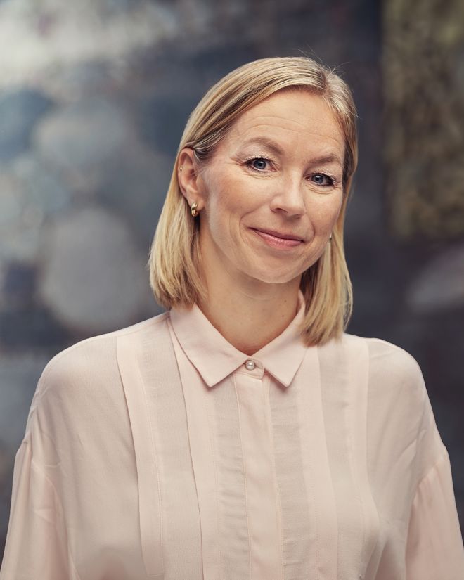 Et portrett av Monica Svenskerud. Hun står med skulderlangt blondt hår og har på seg en lys rosa skjorte med krage. Bakgrunnen er mykt uskarp med duse blå og grå toner, og gir bildet en rolig og nøytral stemning.