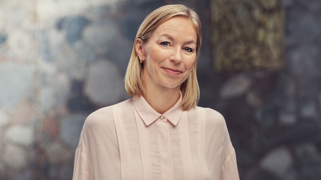 Ei kvinne med lyst, skulderlangt hår står framfor ein uklar bakgrunn og har på seg ein lys skjorte med krage.