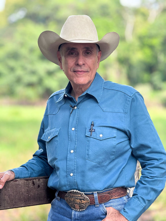 En eldre mann med cowboyhatt og blå skjorte står ved et tregjerde med grønne trær i bakgrunnen.