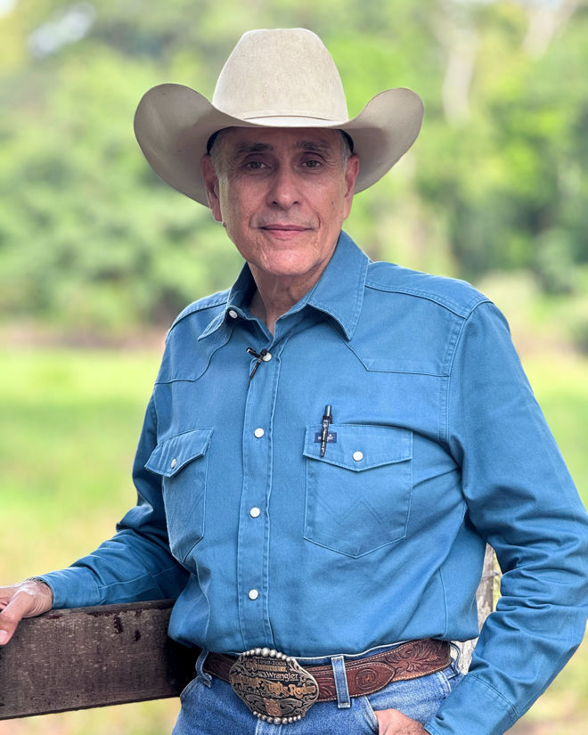 An elderly man wearing a cowboy hat and a blue shirt is standing by a wooden fence with green trees in the background.
