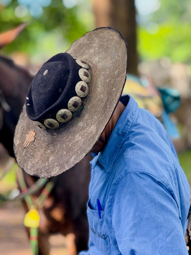 En mann med bred hatt med dekor og blå skjorte står utendørs ved siden av en hest.