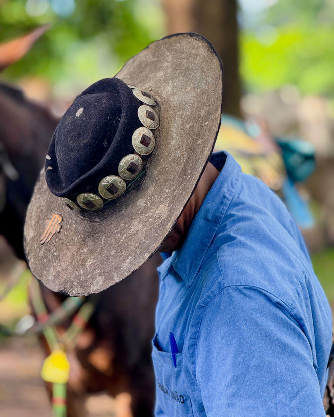 A man wearing a wide decorated hat and a blue shirt is standing outdoors next to a horse.