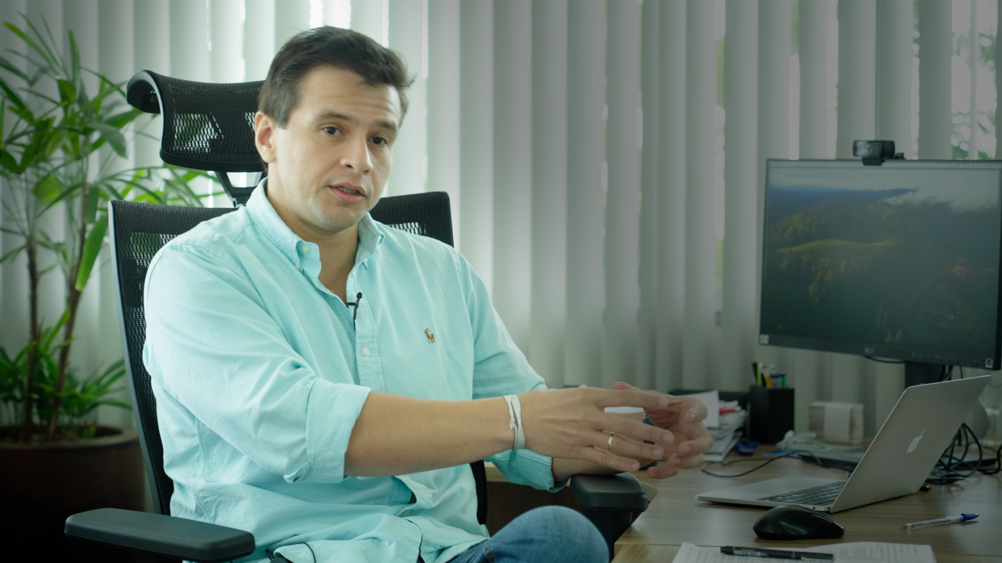 En ung mann med lys skjorte sitter i en kontorstol ved et skrivebord med dataskjerm, bærbar PC og kontorutstyr.