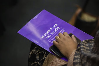 Bildet viser en hånd som hviler på et lilla konferanseprogram med teksten "Women, Peace and Security: The 25th Anniversary Conference of UNSCR 1325, 16 May 2025." Ved siden av programmet ligger en mobiltelefon. 