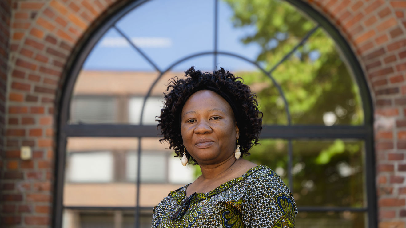 The image portrays a woman standing in front of a large arched window set into a brick wall. The window's design features multiple panes and frames a view of a tree with green leaves and a building in the background