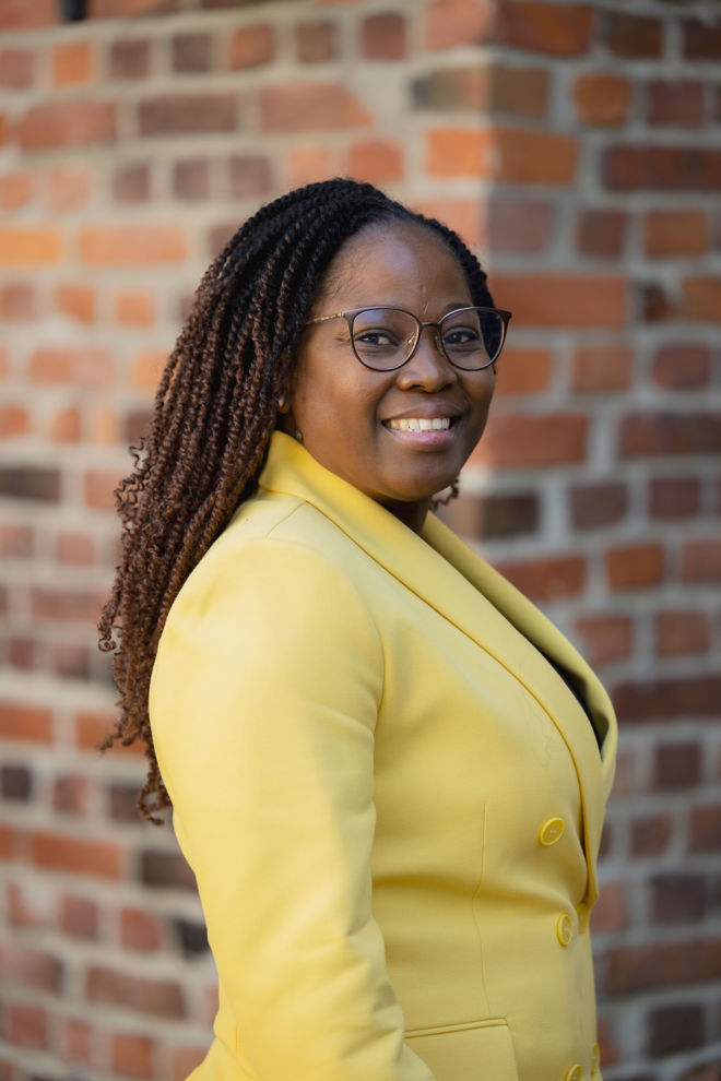 Bildet viser en person med mørkt, flettet hår, kledd i en lys gul blazer med knapper. Bakgrunnen består av en murvegg med røde og oransje mursteinsdetaljer, som gir et urbant og klassisk preg. Den gule blazeren gir en livlig kontrast mot den varme mursteinsbakgrunnen.