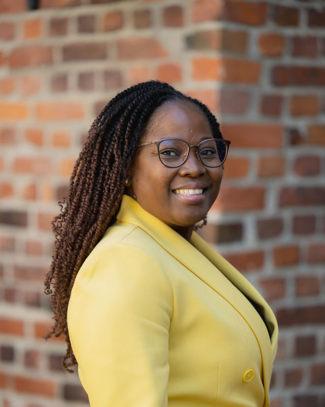 The image features a woman wearing a bright yellow blazer with button details, standing against a brick wall.