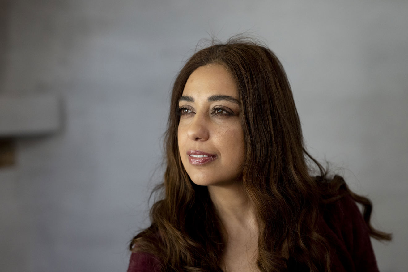 The image shows a woman with long, brown hair, wearing a dark-colored top, set against a neutral gray background. The lighting is soft, creating a calm and subdued atmosphere.