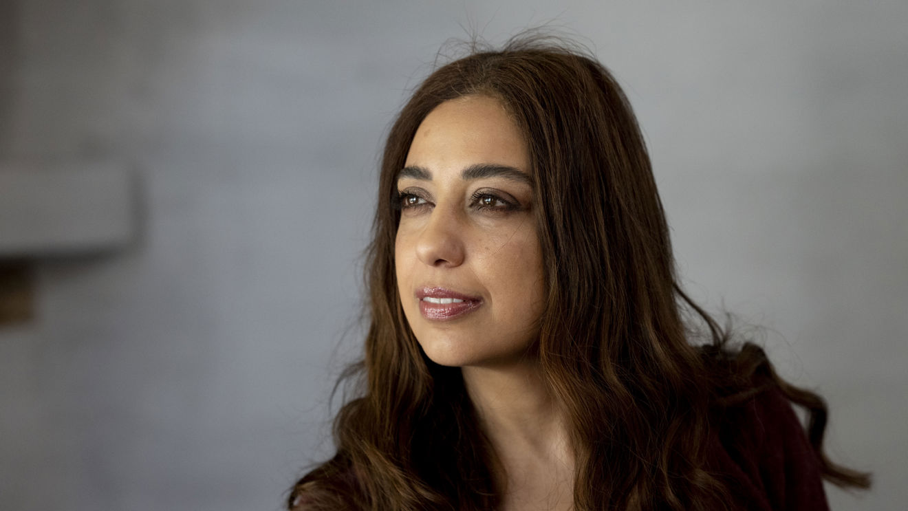 The image shows a woman with long, brown hair, wearing a dark-colored top, set against a neutral gray background. The lighting is soft, creating a calm and subdued atmosphere.