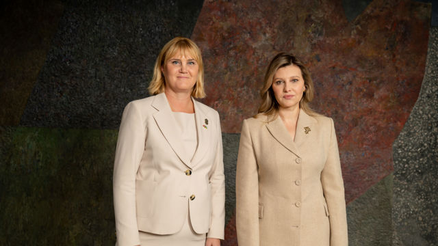 To kvinner står side om side foran en kunstnerisk, fargerik bakgrunn. Begge har på seg lyse drakter med blazer og skjørt eller bukse. Den ene har skulderlangt lyst hår, mens den andre har langt, brunt hår. Begge har en brosje festet på jakkeslaget. De står oppreist og ser rett frem.