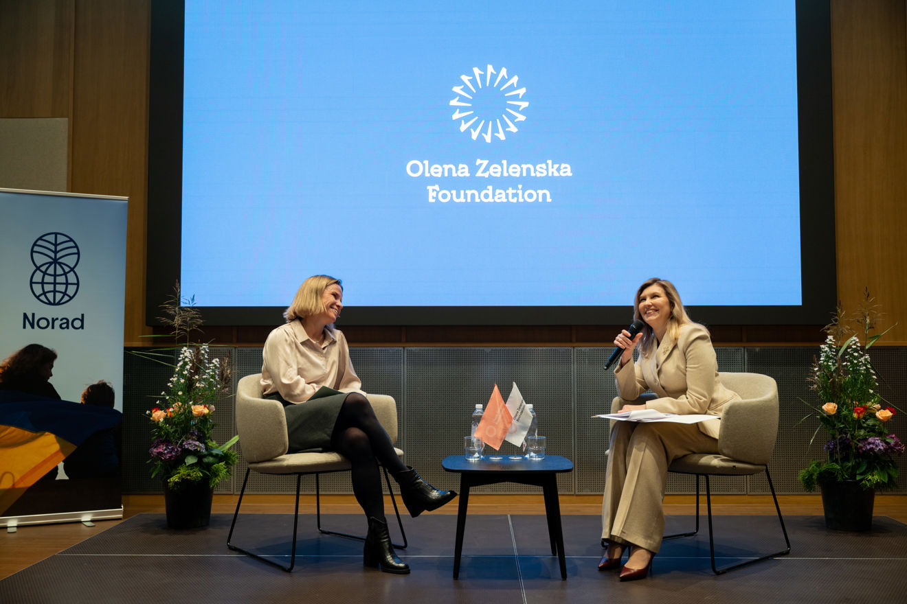 Two women are seated on stage in modern, light-colored chairs, engaging in a conversation. One is holding a microphone and some papers, while the other listens attentively with her legs crossed. Between them is a small table with water bottles and small flags. In the background, a large blue screen displays the text "Olena Zelenska Foundation" along with the foundation's logo. To the left, a banner with the Norad logo is visible, and there are flower arrangements on both sides of the stage. The setting suggests a formal event.