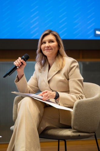Ukrainas førstedame Olena Zelenska sitter i en moderne, lysefarget stol og har på seg en beige dress. Hun holder en mikrofon i den ene hånden og noen papirer eller en mappe i den andre, noe som tyder på at hun deltar i en samtale eller presentasjon. Bakgrunnen viser en stor, blå skjerm. Bildet er tatt under arrangementet. Hun sitter oppmerksomt og engasjert.