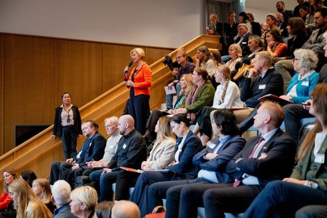 Et auditorium er fylt med mange mennesker som sitter i trapperadene og følger med på et arrangement. En kvinne i oransje jakke står oppreist midt i salen med en mikrofon i hånden, tilsynelatende for å stille et spørsmål eller gjøre et innlegg. Deltakerne er kledd i formelle eller pene klær, og det er en variert gruppe både kvinner og menn. Stemningen er konsentrert og engasjert.