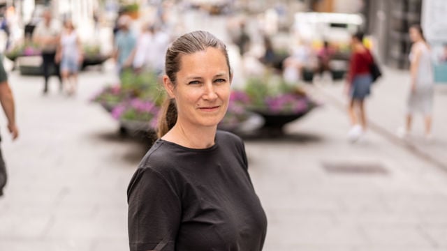 En voksen kvinne med brunt hår i hestehale står ute på en gågate med planter og andre mennesker i bakgrunnen.