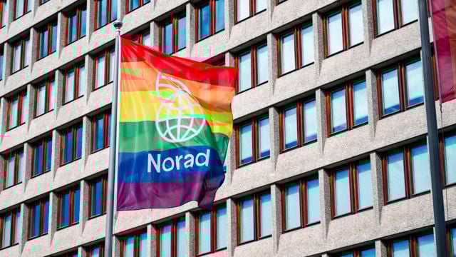 The image depicts a building with many rectangular windows and two rainbow flags flying in front. One flag bears the Norad logo, symbolizing support for LGBTQ+ rights.
