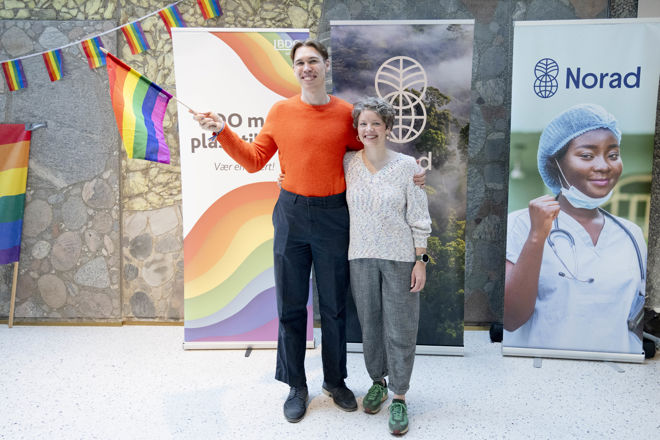 Bildet viser to personer som står sammen foran tre store bannere. Den ene personen holder en regnbueflagg, symbolsk for LGBTQ+-stolthet. 