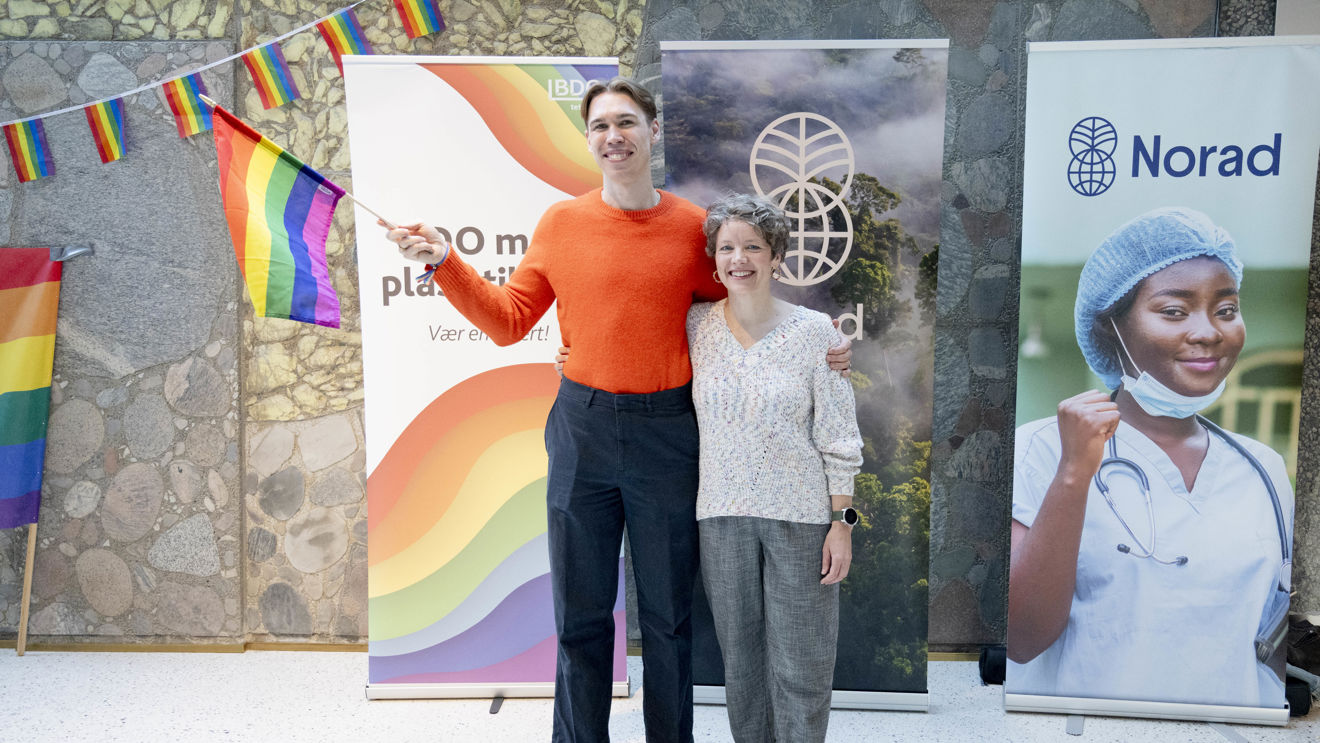 Bildet viser to personer som står sammen foran tre store bannere. Den ene personen holder en regnbueflagg, symbolsk for LGBTQ+-stolthet.