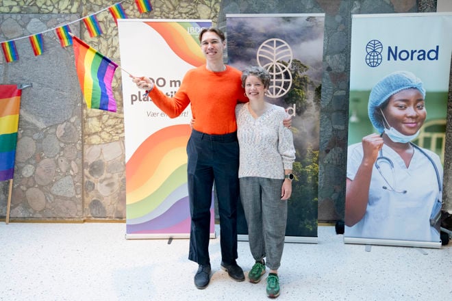 Bildet viser to personer som står sammen foran tre store bannere. Den ene personen holder en regnbueflagg, symbolsk for LGBTQ+-stolthet.