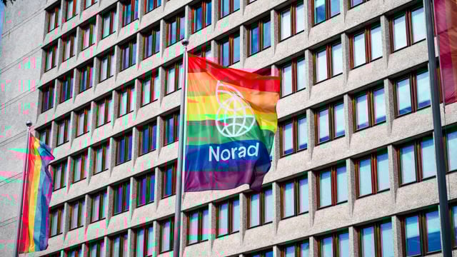 Bildet viser en stor bygning med mange vinduer i bakgrunnen. Foran bygningen vaier to flagg på flaggstenger. Det ene flagget er et regnbueflagg med fargene rød, oransje, gul, grønn, blå, og lilla, og har teksten "Norad" sammen med en stilisert jordklodelogo.