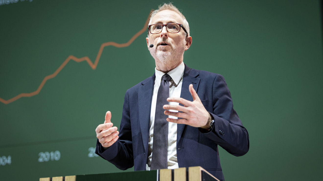 Bård Vegar Solhjell, direktør i Nora, står på en talestol. Grønn tavle i bakgrunnen som viser graf.