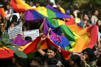 En folkemengde holder opp et stort regnbueflagg under en utendørs markering, med flere plakater synlige i bakgrunnen.