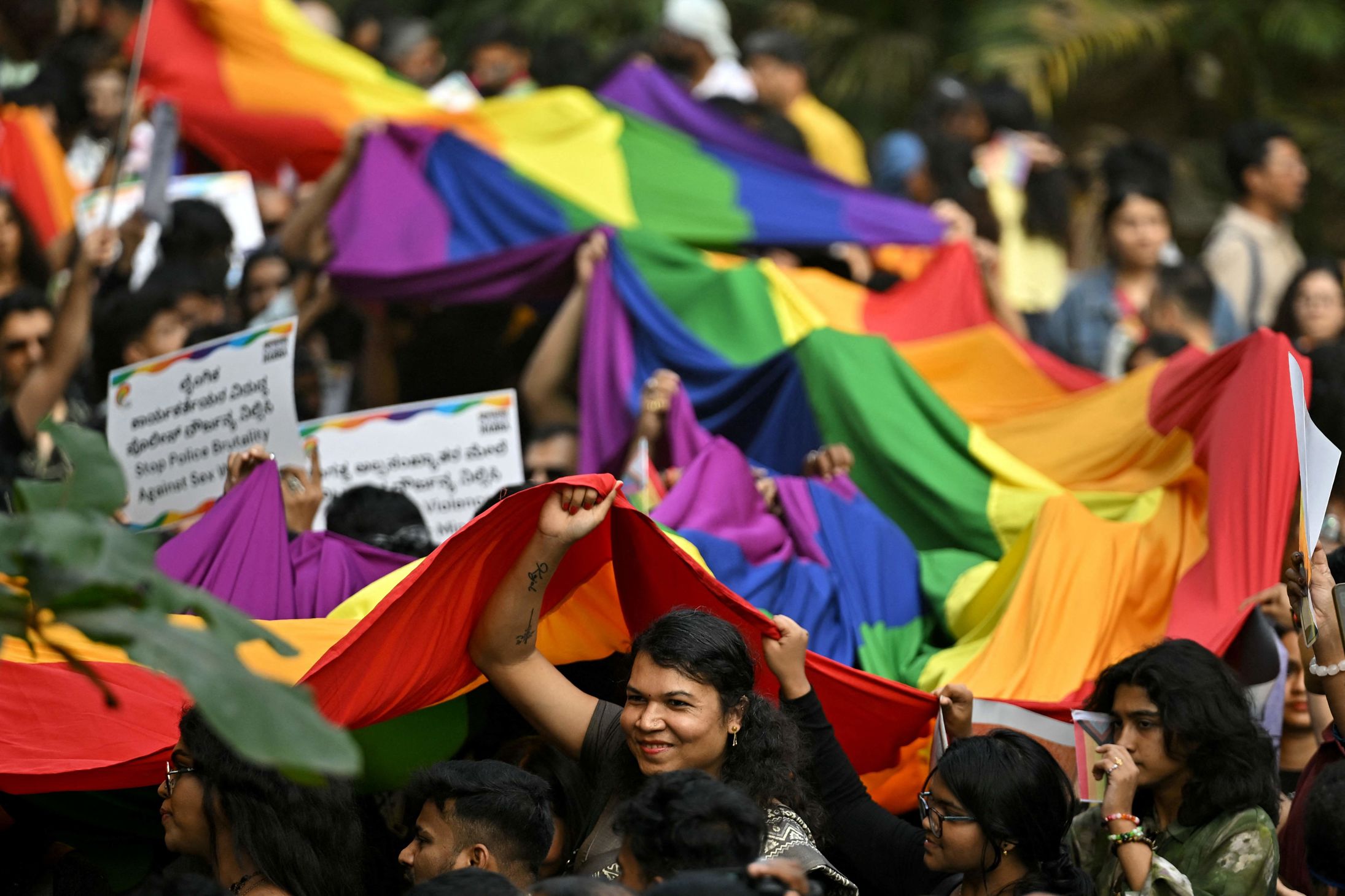 En gruppe mennesker holder opp et stort regnbueflagg under en utendørs parade, med flere plakater synlige i mengden.