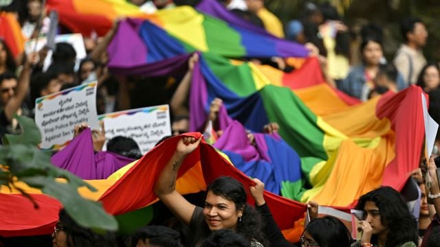 En gruppe mennesker holder opp et stort regnbueflagg under en utendørs parade, med flere plakater synlige i mengden.