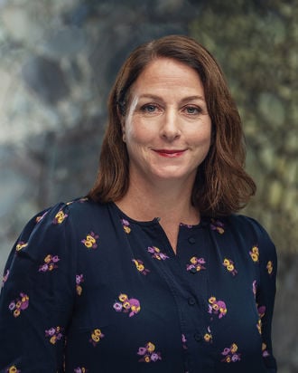 En kvinne med brunt, skulderlangt hår har på seg en mørk bluse med blomster, og står foran en uskarp bakgrunn.