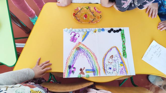 På bildet ser man et gruppebord med flere barn samlet rundt. På bordet ligger et stort ark med en fargerik barnetegning. Tegningen viser to regnbuer, noen blomster, en sommerfugl, en sol, og flere figurer som ser ut som barn.