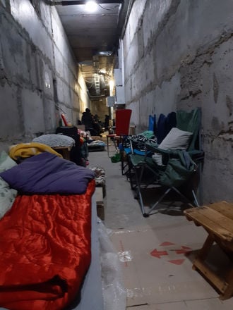 Dette bildet viser en lang, smal gang med betongvegger og et uferdig utseende, som minner om en kjeller eller en tilfluktsbunker. Langs begge sider av gangen står feltsenger, madrasser, soveposer og sammenleggbare stoler. Noen steder ligger det tepper og puter, og det er også små bord og personlige eiendeler plassert rundt. Belysningen er sparsommelig, med et par lyspærer i taket. Litt lenger inn i gangen sitter flere personer samlet, men de er litt uklare og i bakgrunnen.