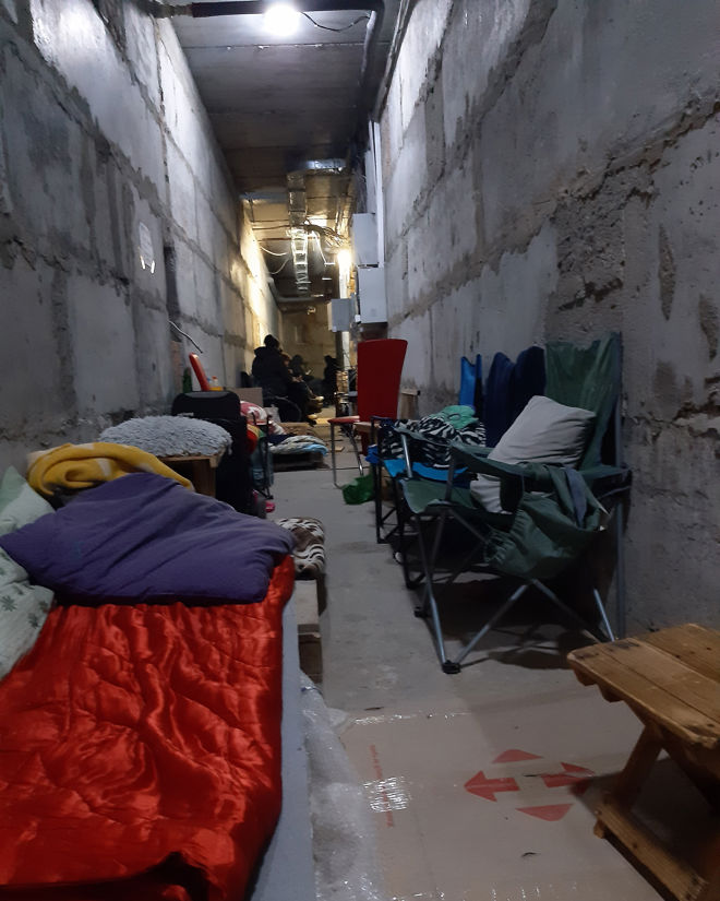 This image shows a long, narrow corridor with concrete walls and an unfinished appearance, resembling a basement or a shelter bunker. Along both sides of the corridor are camp beds, mattresses, sleeping bags, and folding chairs. In some places, there are blankets and pillows, as well as small tables and personal belongings scattered around.