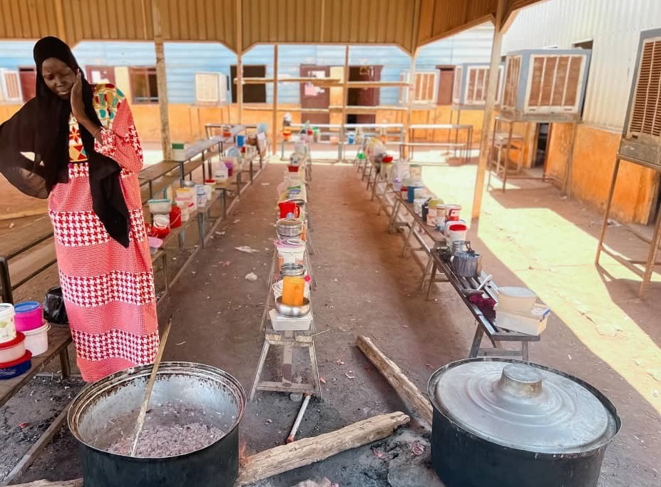 Bildet viser en kvinne i lang, rød- og hvitmønstret kjole og svart sjal som står ved en stor gryte over åpen ild. Gryten står på bakken, og det er en stor tresleiv oppi. Rundt henne er det benker og bord med mange fargerike plastbokser og kjeler, som trolig inneholder mat eller ingredienser.