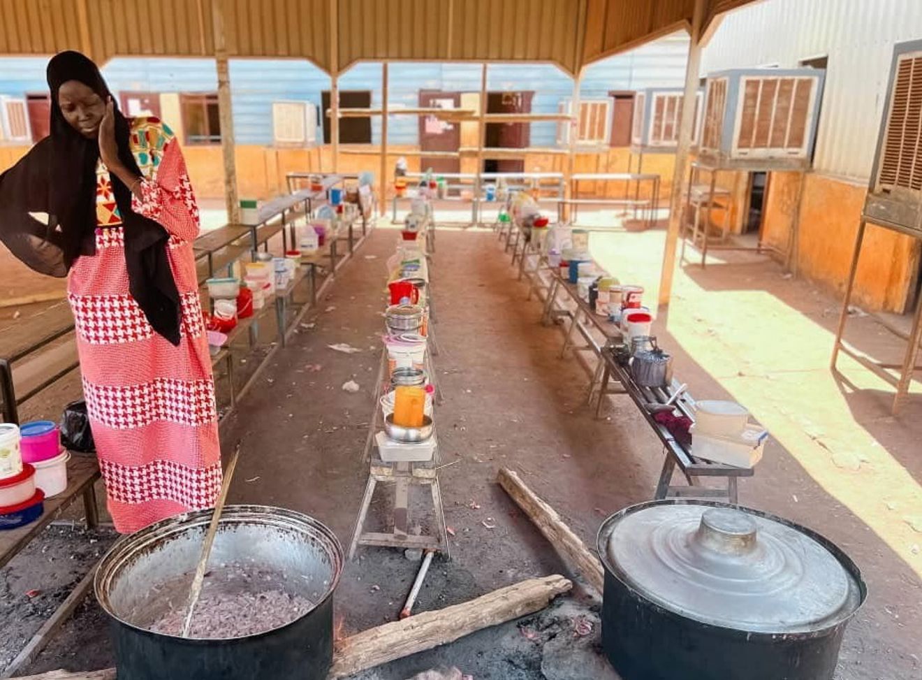 Bildet viser en kvinne i lang, rød- og hvitmønstret kjole og svart sjal som står ved en stor gryte over åpen ild. Gryten står på bakken, og det er en stor tresleiv oppi. Rundt henne er det benker og bord med mange fargerike plastbokser og kjeler, som trolig inneholder mat eller ingredienser.